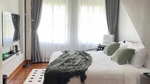Une chambre moderne avec un grand lit, un téléviseur à écran plat et une vue sur l’extérieur à travers des rideaux transparents.