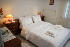 A neatly made bed with white linens and two pillows, a bedside table with a lamp, and a radiator on the wall. - Avli Guesthouse (Trikala)