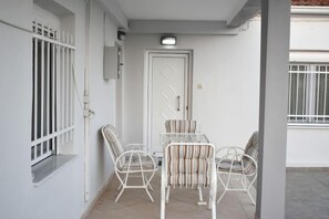 A small patio area with a white door, a table with chairs, and a window with white bars. - Avli Guesthouse (Trikala)