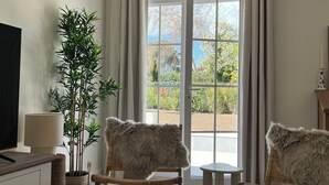 Ein Zimmer mit Fernseher, Blumentopf, zwei Sessel mit Felldecken, einem kleinen Tisch und einem großen Fenster mit Vorhängen.
