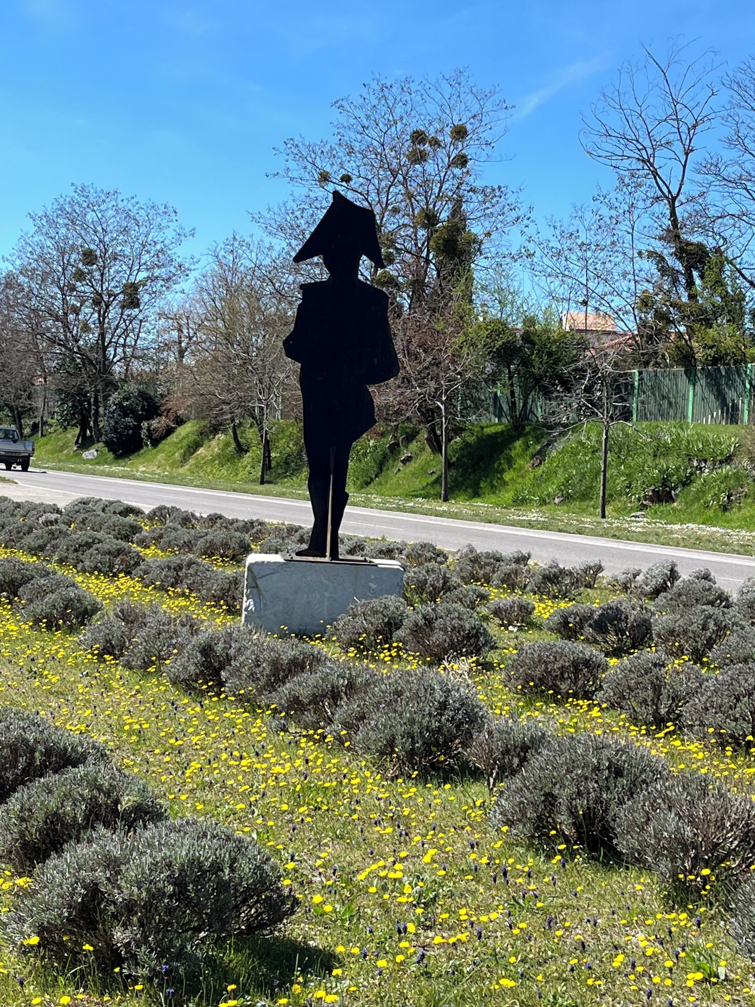 A statue of a woman in a hat standing on a pedestal in a landscaped area with yellow flowers and shrubs.