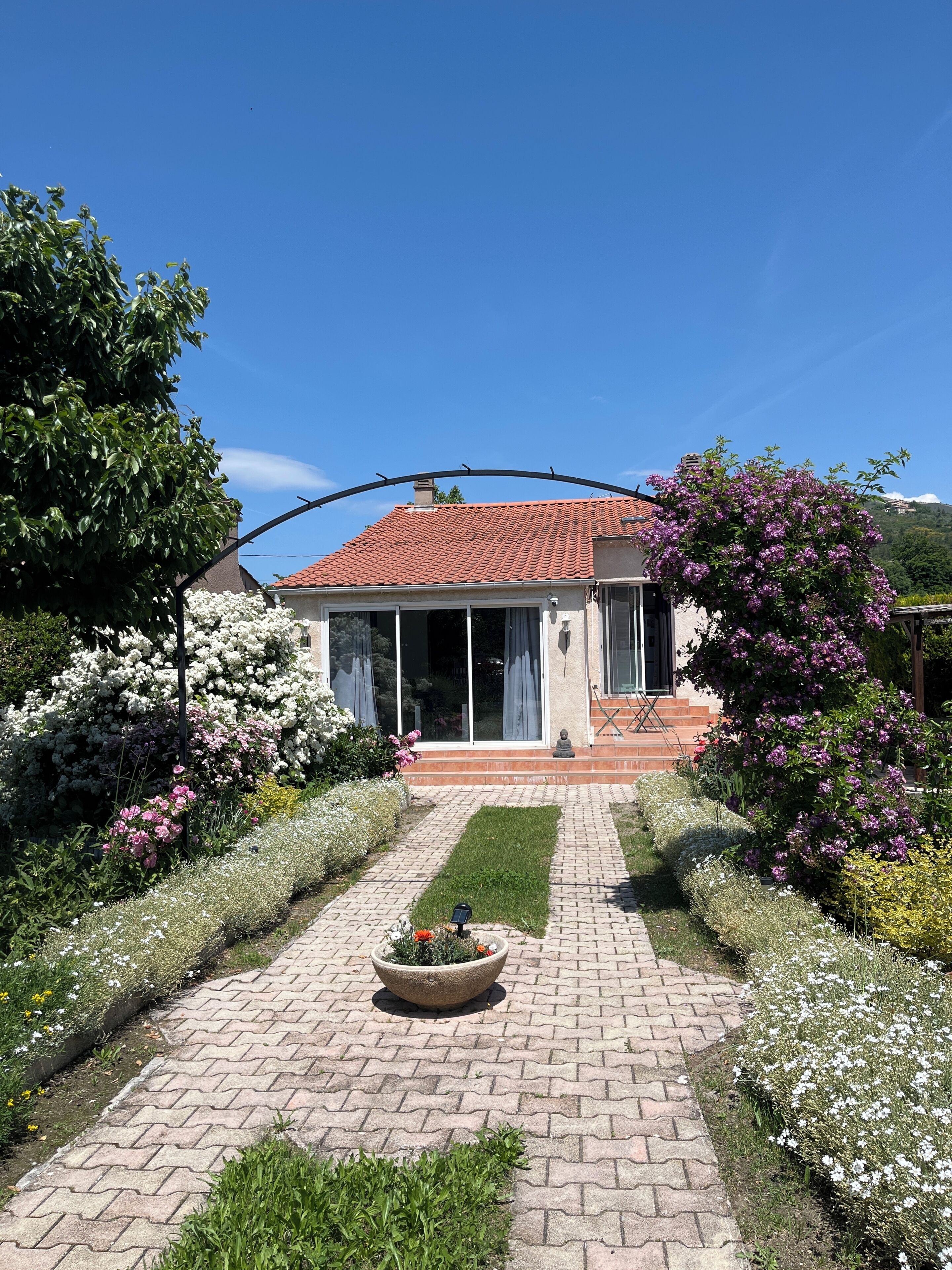 A house with a red-tiled roof, a garden with various plants and flowers, and a paved pathway leading to the entrance.