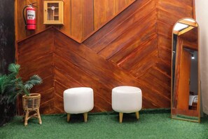 Una habitación con pared de madera en patrón espiga, dos taburetes blancos acolchados, una pequeña planta de mimbre y un extintor de incendios montado en la pared.