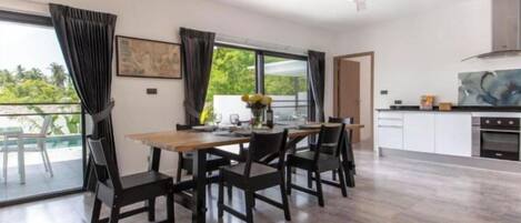 A modern dining area with a wooden table and chairs, a kitchen in the background, and a view of greenery outside.