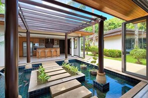 A modern indoor pool area with a pergola, a kitchen in the background, and a view of the outdoors. - Baan Benjamart (Koh Samui)