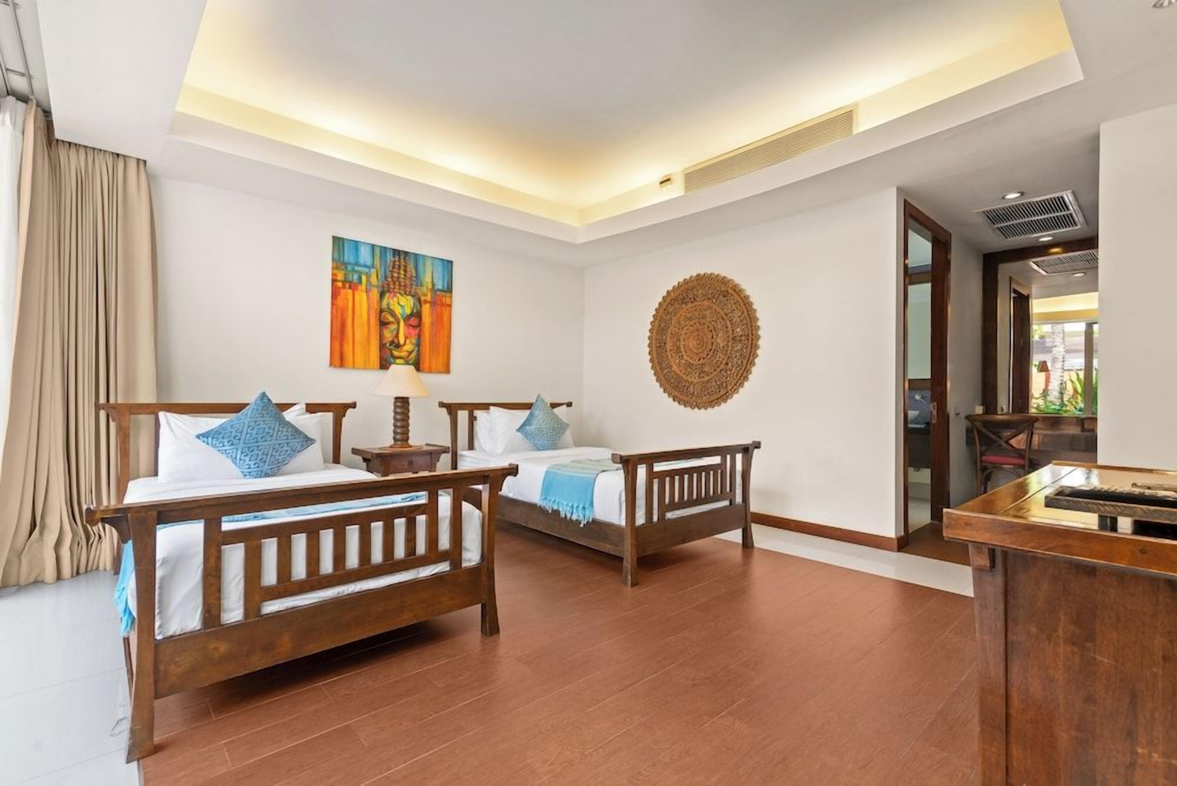 Quarto de hotel com duas camas de madeira, um ornamento circular feito de vime na parede e um quadro.