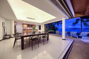 A modern dining area with a large table and chairs, a kitchen in the background, and a view of the ocean at dusk. - Baan Benjamart (Koh Samui)