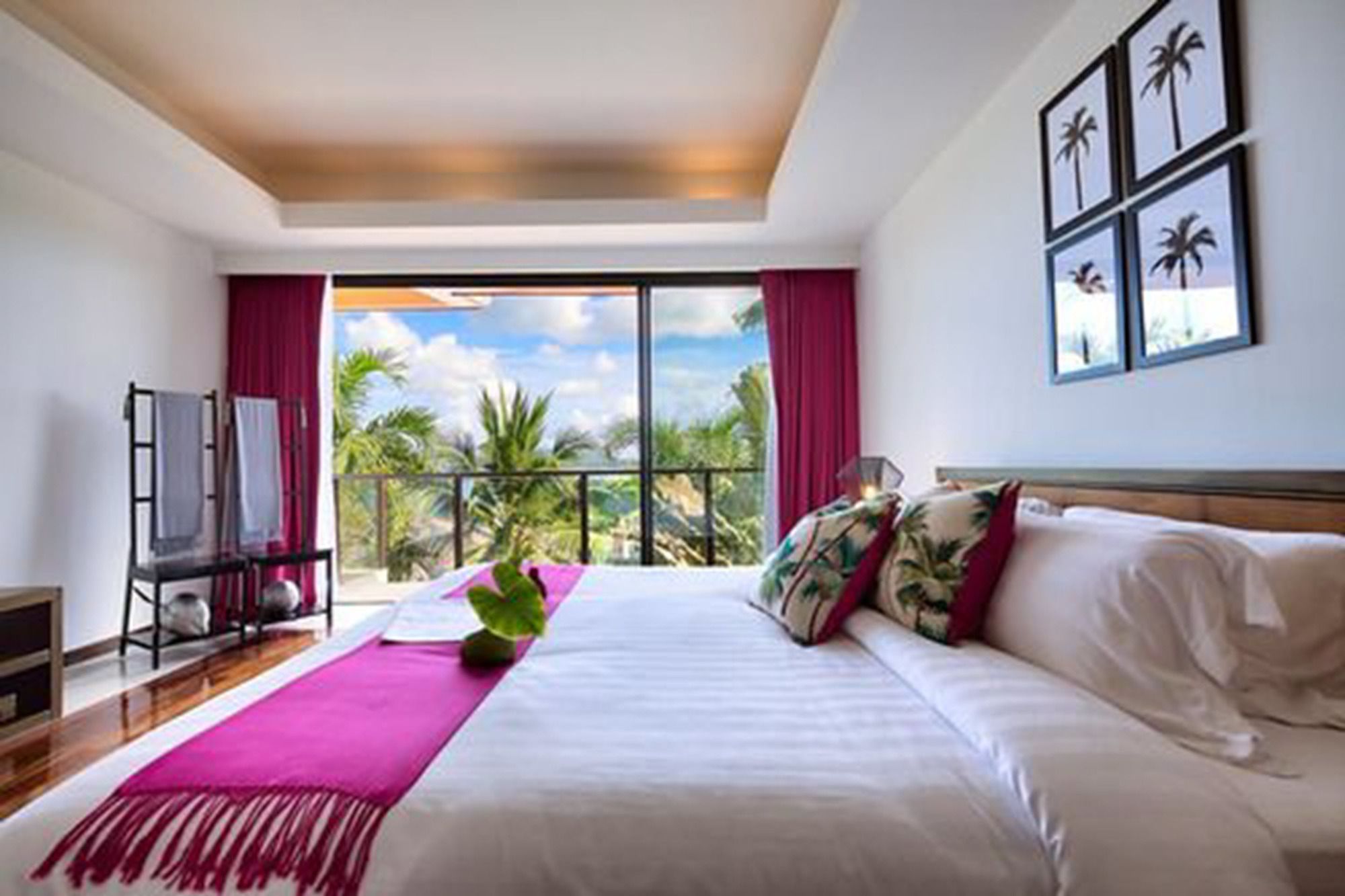 A hotel room with a large bed, a view of palm trees, and a balcony with a red curtain.