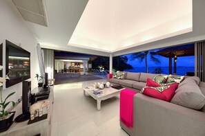 A modern living room with a large sofa, a coffee table, and a flat-screen TV. There is a view of the ocean through large windows. - Baan Benjamart (Koh Samui)