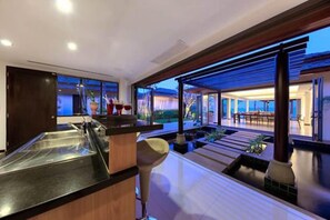 A modern kitchen with a bar area, large windows, and a view of the outdoor seating area. - Baan Benjamart (Koh Samui)
