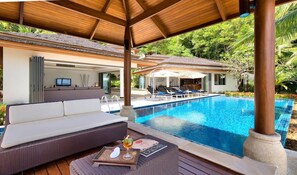 A poolside area with a sofa, lounge chairs, and a table with a drink. - Baan Benjamart (Koh Samui)