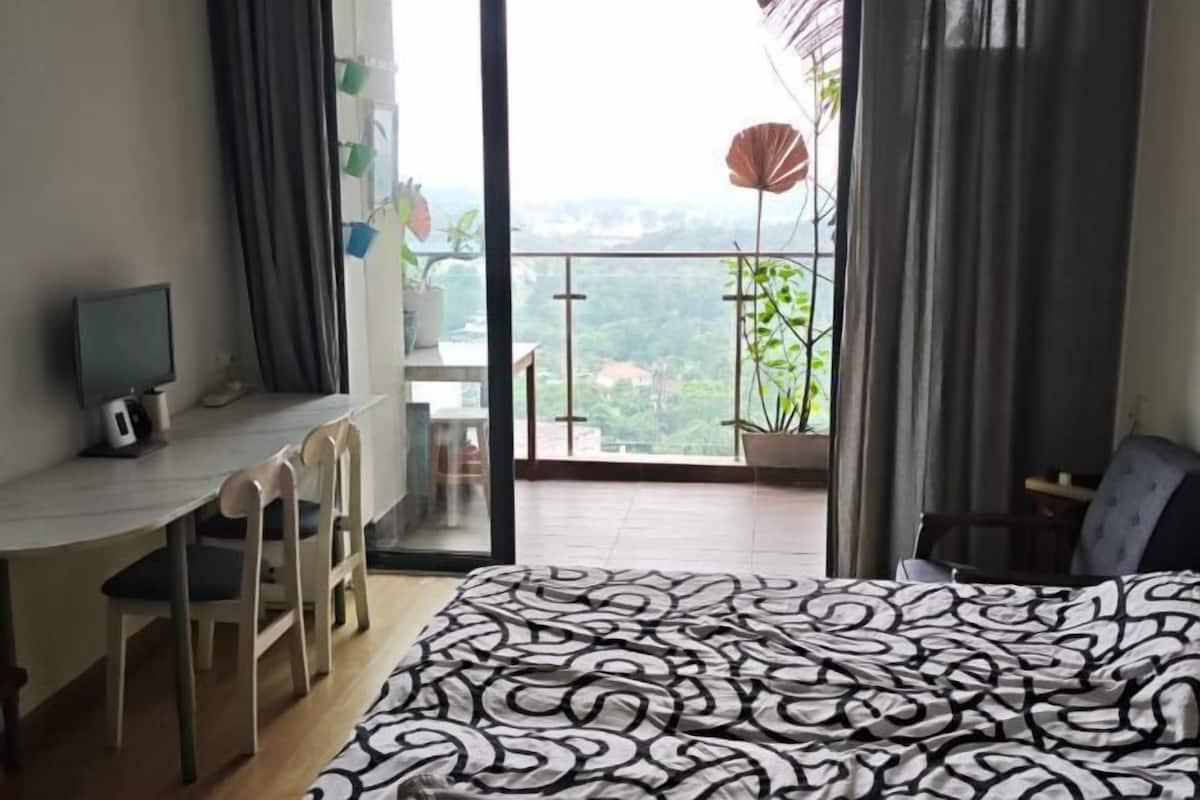 Un dormitorio con cama, escritorio y silla. Se ve una ventana con vista a zonas verdes y edificios.