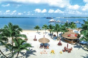 Ein Strand mit Palmen, einer Bar mit Reetdach und Booten im Hintergrund.