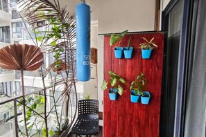 Ein Balkon mit roter Tür, Blumentöpfen, einem schwarzen Stuhl und einem blauen zylindrischen Gegenstand.
