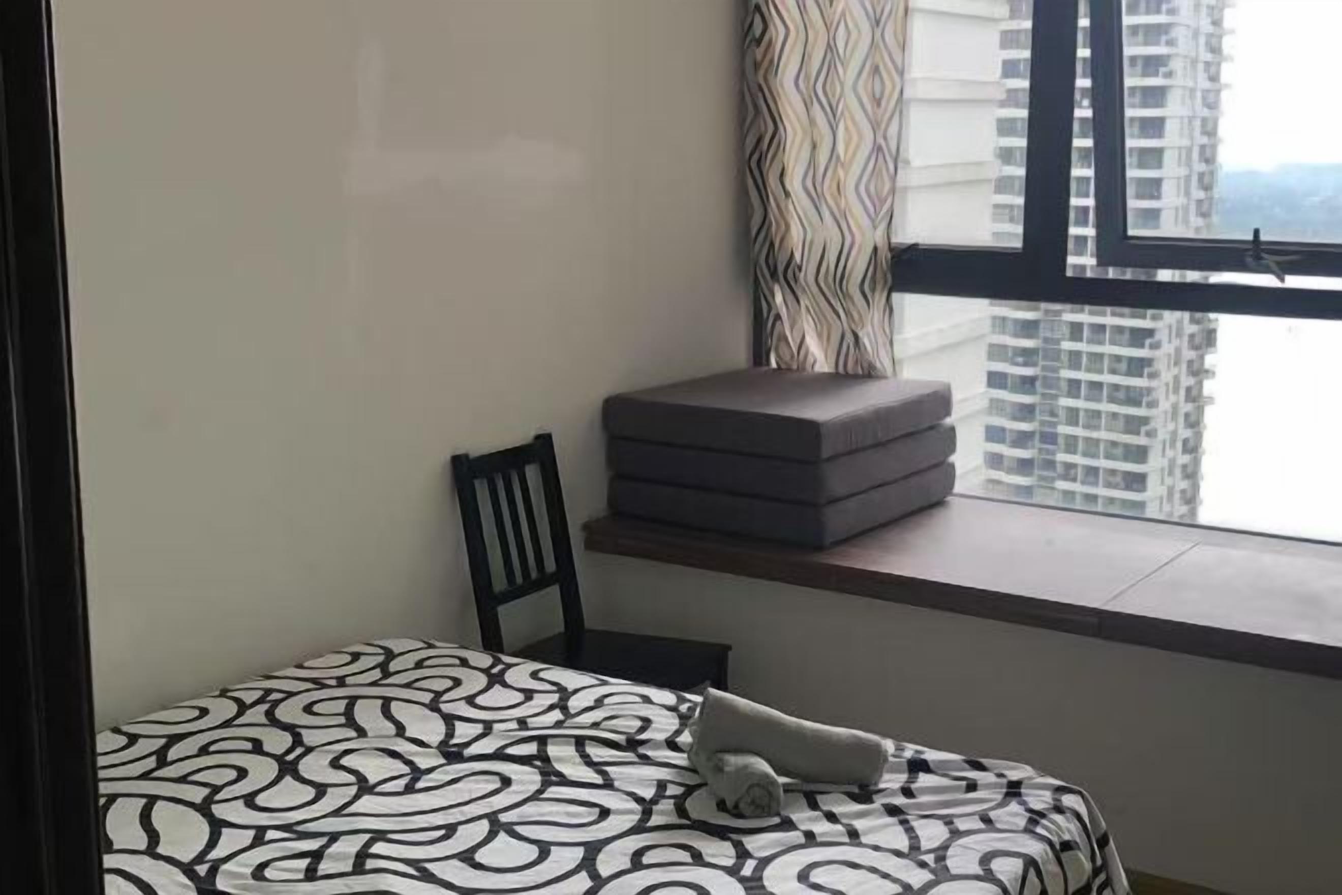 Una camera da letto con una coperta a motivi, una sedia, una pila di cuscini e una finestra con vista su edifici alti.