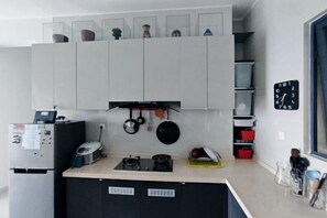 Eine Küche mit weißen Schränken, einem Herd und hängenden Töpfen. An der Wand hängt eine Uhr und zur Rechten befindet sich ein Fenster.