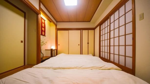 A traditional Japanese-style room with tatami flooring, a low bed with a white bedspread, sliding doors, and a ceiling light. - Ryokan -Ryo- by Tranova (Tokyo)