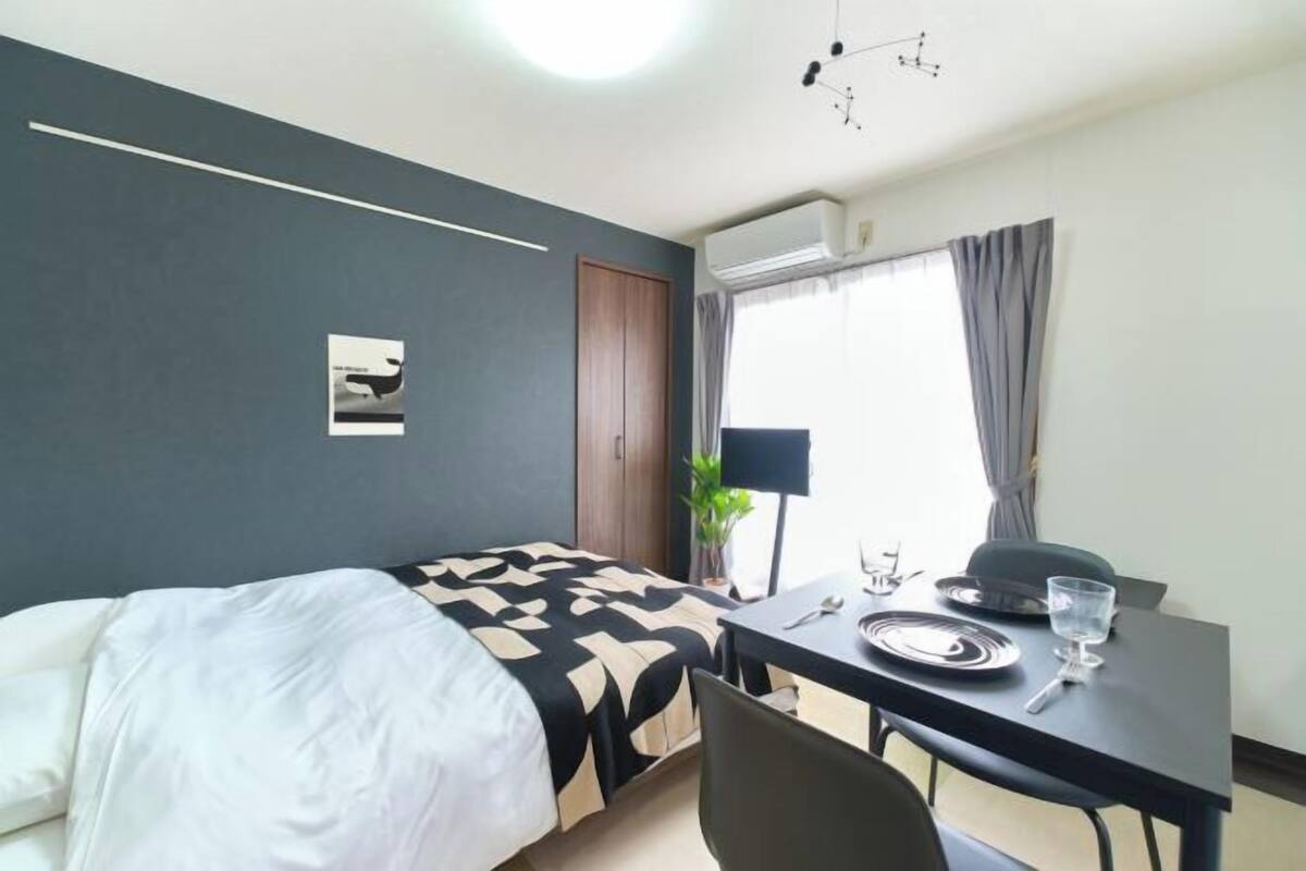A hotel room with a bed, a dining table with plates and glasses, a potted plant, and a window with curtains.