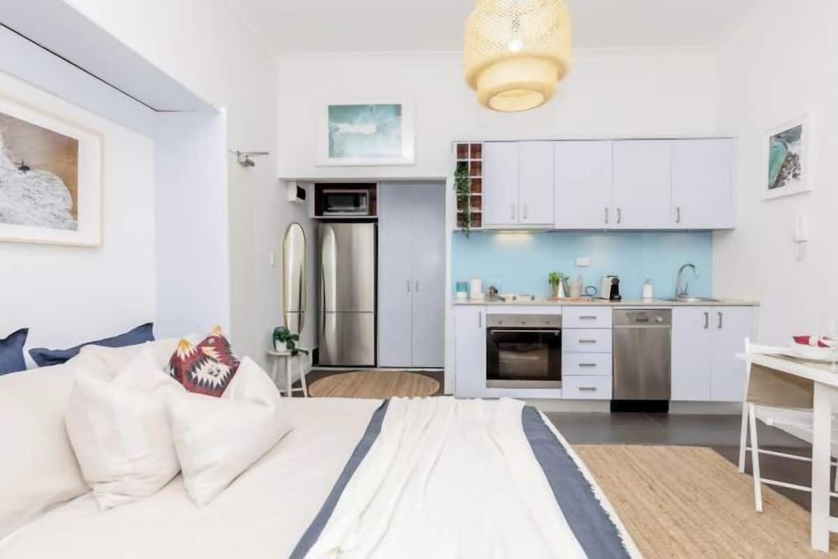 A modern bedroom with a bed, bedside table, and a view of a compact kitchen with cabinets and appliances.