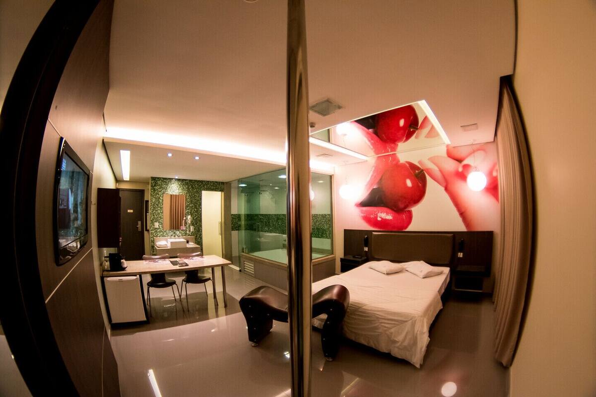A modern hotel room with a large bed, a desk with a chair, a small table, a TV, and a bathroom with a shower and a mirror.