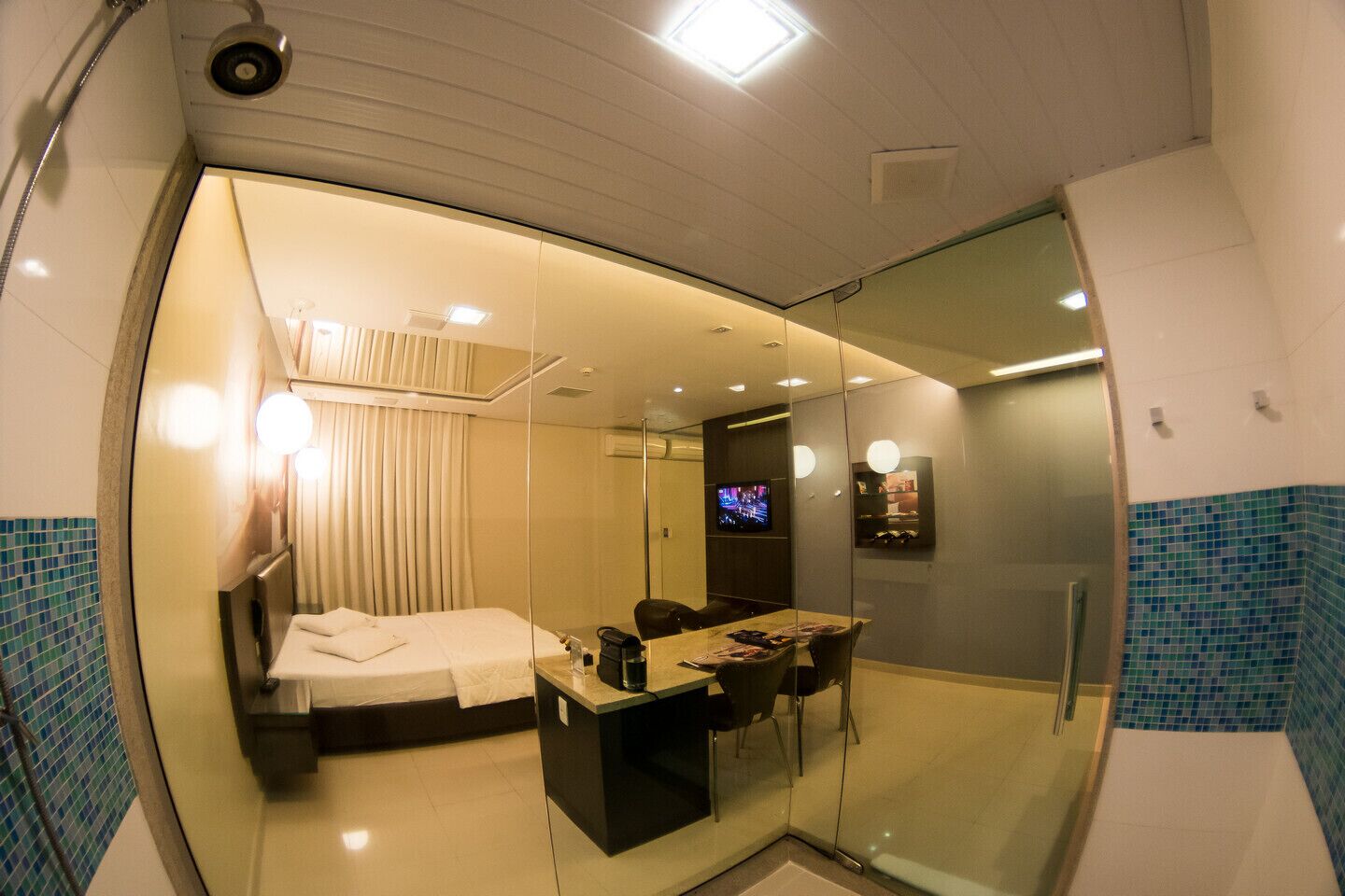 Una habitación de hotel moderna con un separador de vidrio que divide el área de dormir y el baño. La habitación cuenta con una cama, un escritorio con silla, un televisor y una pequeña zona de estar.