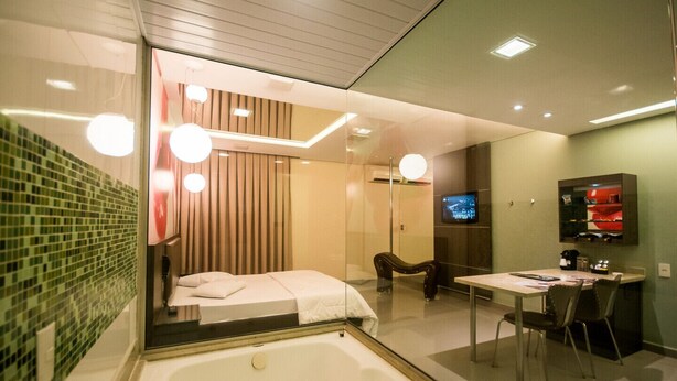 A modern bathroom with a glass shower enclosure, a bathtub, a vanity with a mirror, and a television mounted on the wall. - Motel Vitara (Taboão da Serra)