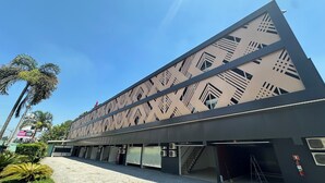 A modern building with geometric patterns on the facade, a covered walkway, and palm trees in the foreground. - Motel Vitara (Taboão da Serra)