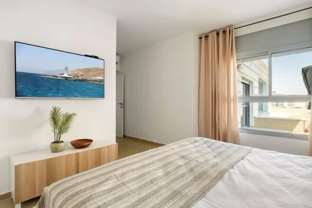 Une chambre d’hôtel moderne, équipée d’un lit, d’un téléviseur fixé au mur, d’une petite plante et d’une fenêtre avec des rideaux.