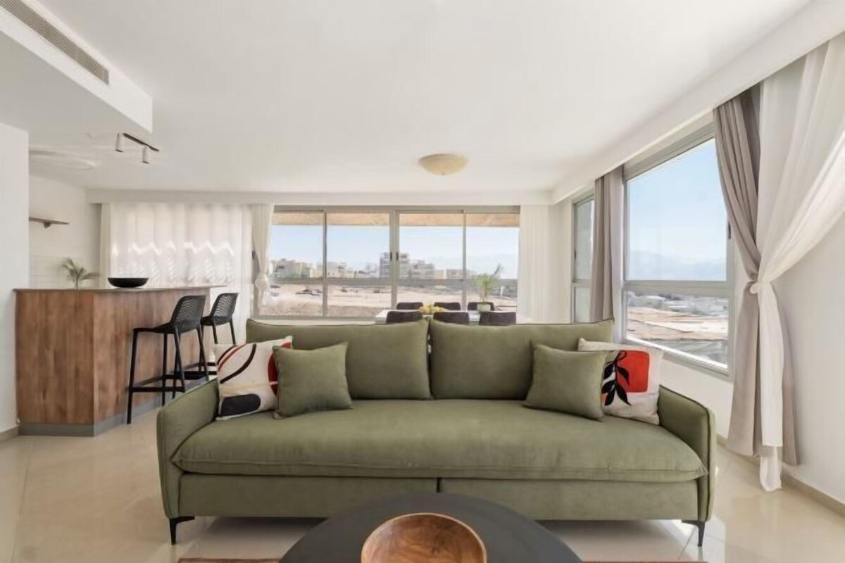 A modern living room with a green sofa, a wooden coffee table, and a view of the city through large windows.