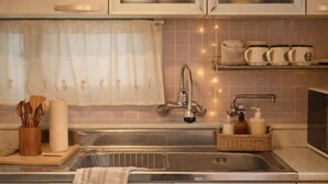 A kitchen with a stainless steel sink, a wooden utensil holder, a roll of paper towels, a wicker basket with toiletries, and a window with a floral curtain. - Turtle Stay Seoul-- the Center of Seoul #Cozy Apartment (Seoul)