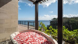 A hot tub filled with red petals, a balcony with a view of the sea, and a stone wall. - By The Cliff Bali Villa (Jimbaran)