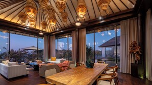 A spacious dining area with a long wooden table and chairs, surrounded by large windows offering a view of the cityscape. - By The Cliff Bali Villa (Jimbaran)