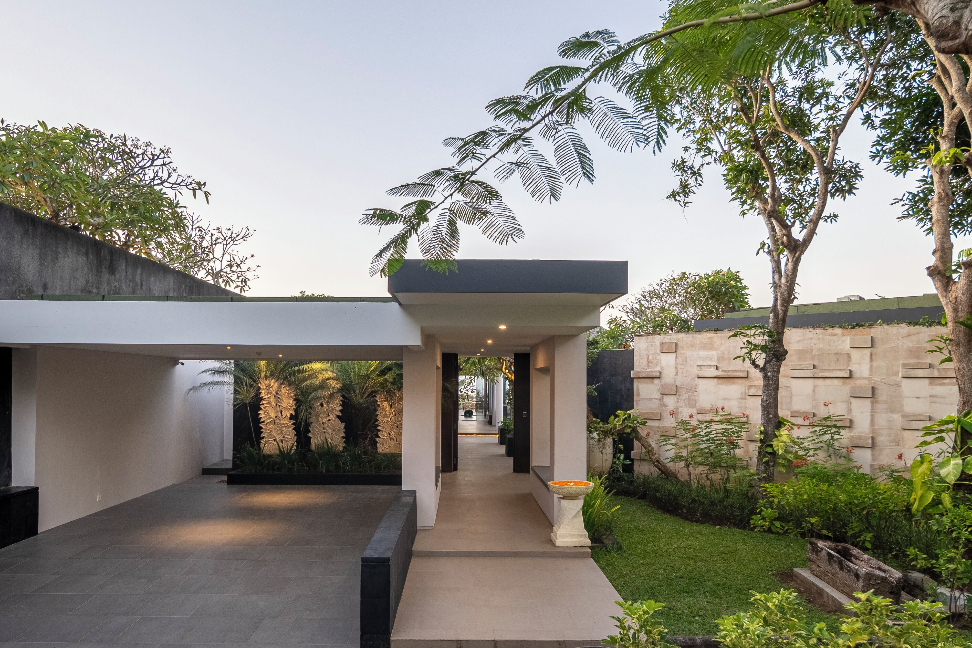 A modern outdoor space with a pathway leading to a covered area, surrounded by greenery and a stone wall.