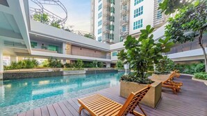Un espace piscine extérieur moderne, doté de chaises longues en bois et de jardinières, entouré de verdure et situé à proximité d’immeubles de grande hauteur.