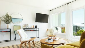 Ein modernes Wohnzimmer mit Sofa, Sessel und Couchtisch, einem flachbildfernseher an der Wand und einem großen Fenster mit Blick ins Freie.