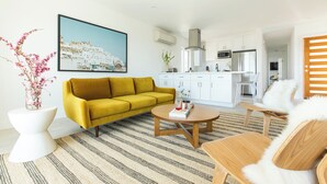 Ein modernes Wohnzimmer mit einem gelben Sofa, einem runden Couchtisch und einem weißen Beistelltisch mit Blumen. An der Wand hängt ein großes Gemälde einer Küstenstadt, und das Zimmer geht in eine Küche mit weißen Schränken und einem Edelstahl-Kühlschrank über.