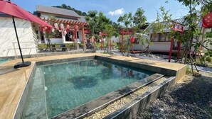 Una piscina con bordeado de baldosas, rodeada de un jardín con árboles pequeños y una pérgola.