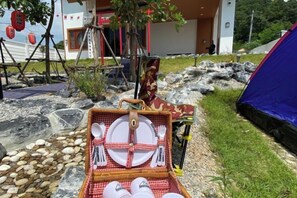 A picnic setup with a basket containing plates, bowls, and cups, a red and white checkered tablecloth, and a blue tent in the background. - Taka Onzen (Kaeng Khoi)
