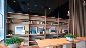 A modern interior with wooden shelves displaying framed pictures and books, a central wooden table, and potted plants.
