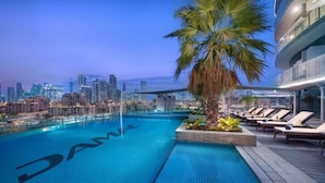 A rooftop pool with a city skyline view, lounge chairs, and a palm tree. - Dazzling Downtown Retreat w Panoramic City Views (Dubai)