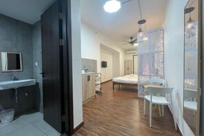 A modern hotel room with a wooden floor, a bed, a desk, and a chair. There is a bathroom with a sink and a mirror. - Urban Retreat @ K Avenue • Pool • Paid Parking (Donggongon)