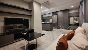 Un salon moderne avec un canapé, une table basse et un téléviseur. On aperçoit à l’arrière-plan un coin cuisine avec un réfrigérateur et une table à manger.
