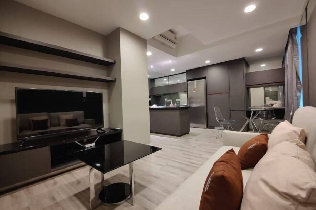 Een moderne woonkamer met een bank, een salontafel en een televisie. Achtergrond toont een keuken met een koelkast en een eettafel.