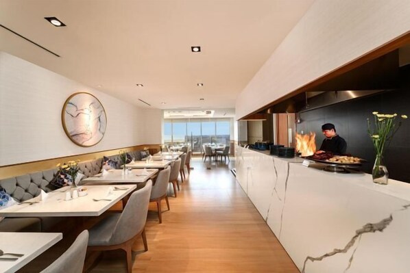 A modern restaurant interior with a long counter, seating area, and a chef preparing food. - Gold Coast Pik Atlantic Sea View Apartment Minimum 2 Nights Stay (North Jakarta)