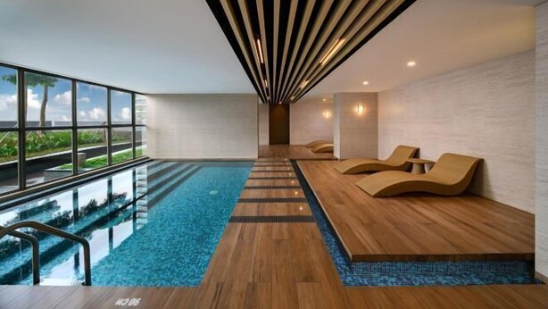 A modern indoor pool area with a clear view of the sky and greenery outside, wooden flooring, and lounge chairs. - Gold Coast Pik Atlantic Sea View Apartment Minimum 2 Nights Stay (North Jakarta)