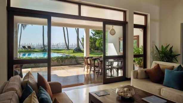 A modern living room with a large sliding glass door leading to a pool area, a sofa with cushions, a coffee table, and a hanging light fixture. - Ranna Beach House - Your own haven by the beach (Hambantota)
