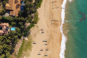 Une vue aérienne d’une plage, avec une rangée de cabanes de plage, une démarcation nette de la ligne de rivage et un filet de pêche visible.