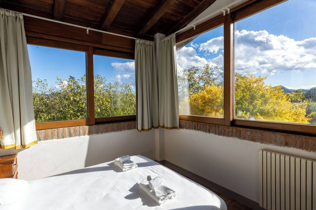 A bedroom with a large bed, two pillows, and a view of trees and mountains through the windows.