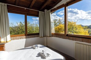Quarto com cama grande, duas almofadas e vista para árvores e montanhas pelas janelas.