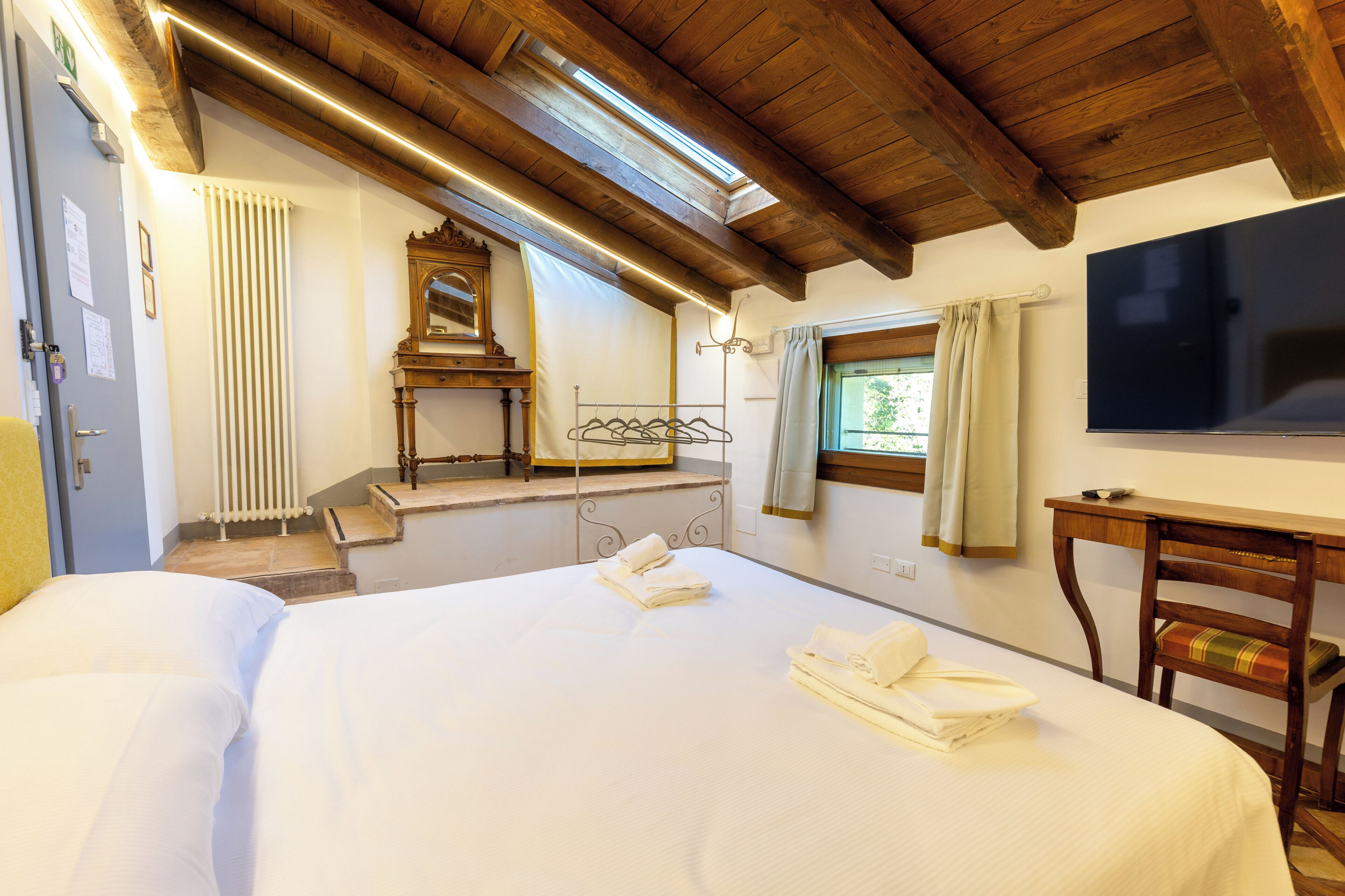Quarto de hotel com uma cama grande, teto de madeira, televisão, janela com cortinas e radiador.